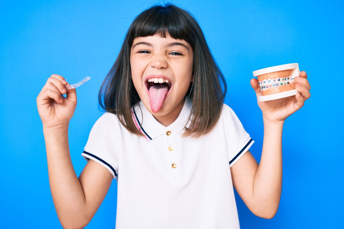 Niña con flequillo sosteniendo alineadores invisibles y brackets, sacando la lengua, feliz y con una expresión divertida.