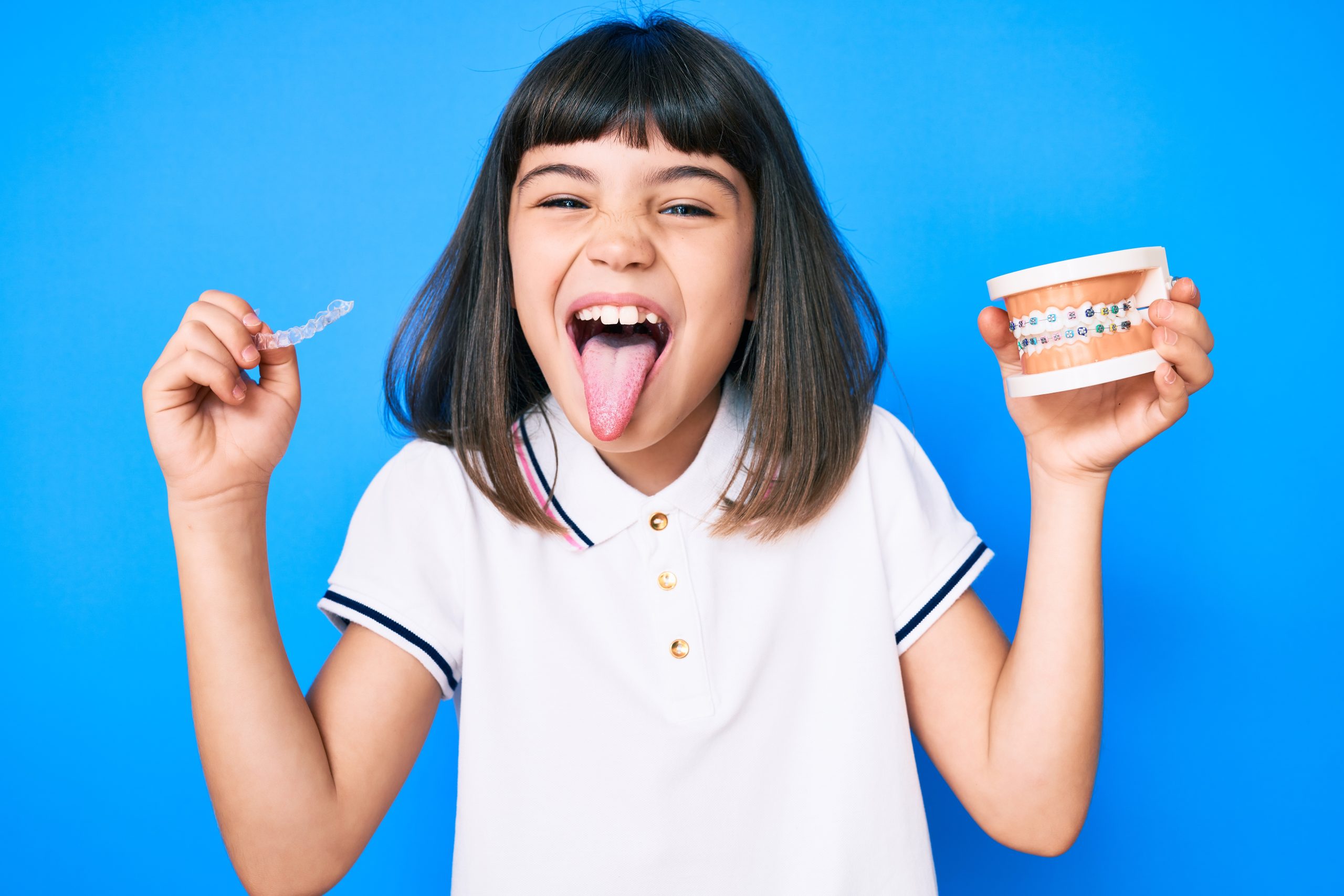 Niña pequeña con flequillo sosteniendo alineadores invisibles y brackets, sacando la lengua, feliz y con una expresión divertida.